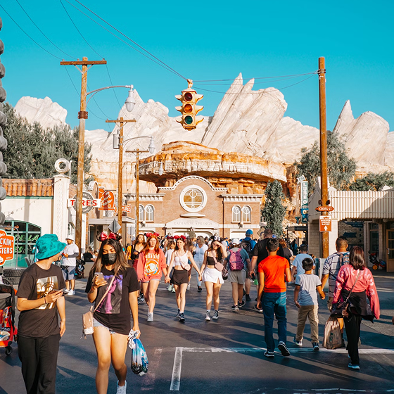 A crowded pedestrian street at a theme park, featuring a central brick building beneath a yellow traffic light, with towering, sculpted rock formations in the background.