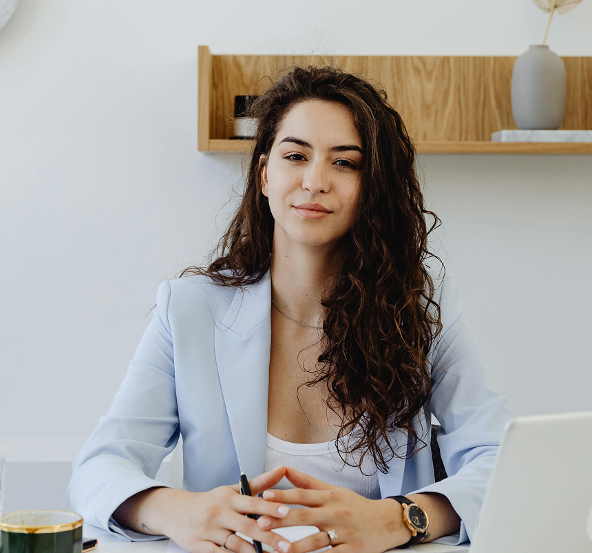 A woman with long dark curly hair, wearing a light blue blazer, sits at a white desk and looks at the camera with her hands clasped in front of a laptop.